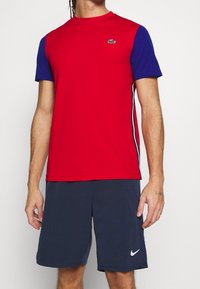 Man med en röd och blå kortärmad skjorta med en liten krokodil-logotyp och marinblå shorts med en vit Nike-logotyp.