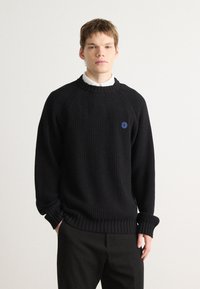 Pulover negru din tricot cu model cu guler rotund, având un logo al unui zâmbet albastru pe piept și mâneci lejere, purtat împreună cu pantaloni.