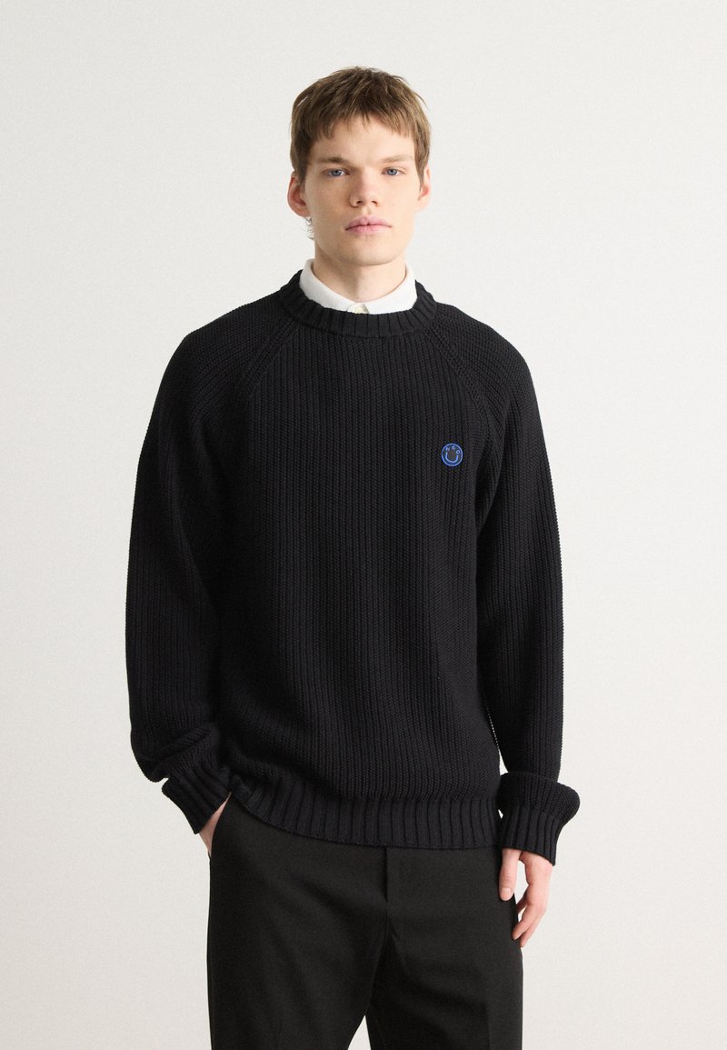 Pulover negru din tricot cu model cu guler rotund, având un logo al unui zâmbet albastru pe piept și mâneci lejere, purtat împreună cu pantaloni.