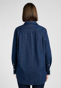 Mörkblå denimskjorta med krage, långa ärmar och knäppta manschetter. Har en avslappnad passform med en jämn textur och subtil lyster.