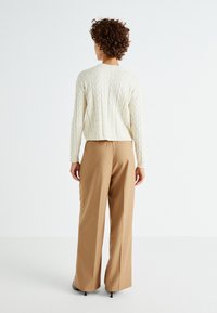 Pull en maille torsadée crémeux à manches longues, associé à un pantalon large de couleur beige clair. Le look est complété par des chaussures à talons lisses.