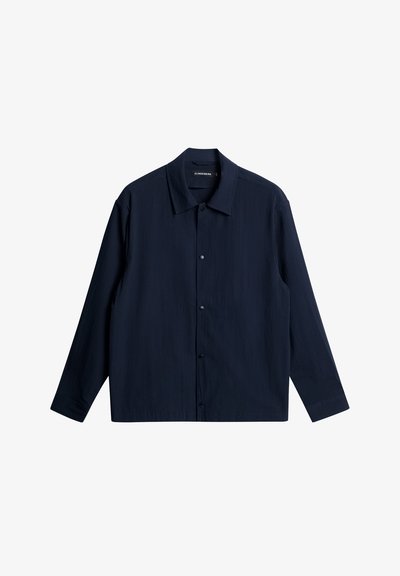 Tummansininen pitkähihaine paita, joka on valmistettu teksturoidusta kankaasta. Paidassa on levitetty kaulus, etunapit ja suora helma.