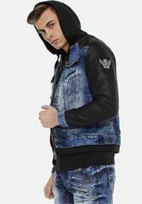 Denim jacket with black leather sleeves, blue faded wash, zip closure, and embroidered patch on the left arm. Black hoodie underneath.
