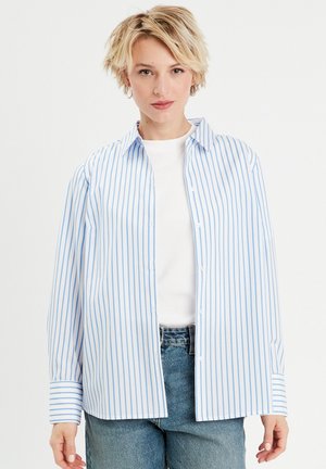 Chemise à rayures bleues claires et blanches avec col, manches longues et coupe décontractée, associée à un haut blanc et un jean en denim.