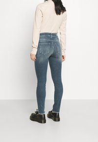 Jeans de tiro alto de color azul con un ajuste Slim, que presentan dos bolsillos traseros y una textura desgastada. Combinados con zapatos negros, vistos desde atrás.