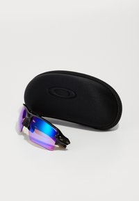 Óculos de sol com armação preta e lentes em degradê azul-rosa, colocados ao lado de uma caixa dura oval preta, com superfície texturizada e logótipo.