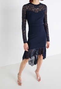 Kvinna iklädd en marinblå klänning med långa spetsärmar, spetsdekoration vid yoken och asymmetrisk spetskant, tillsammans med silverfärgade sandaletter med remmar.