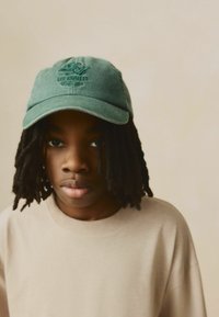 Gorra de algodón verde con el logotipo bordado "LA LOS ÁNGELES". El portador lleva un sudadera beige y tiene el pelo oscuro y texturizado.