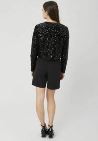 Sweat-shirt noir orné de sequins, avec un col rond, associé à un short noir sur mesure et des sandales à talons noires. Vue de dos.