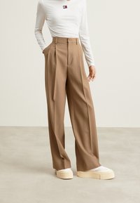 Wide-leg, beige trousers with front pleats, high waist, and side pockets. Paired with white top and chunky, textured-soled sneakers.