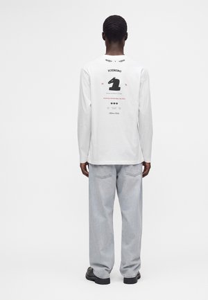 Mand iført hvid langærmet skjorte med sort ridder skakbrik grafik og tekst på ryggen, lyseblå jeans og sorte sko, stående med ryggen til.