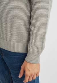 Maglione grigio lavorato a maglia con un motivo testurizzato, polsini e orlo a coste. Indossato sopra dei jeans blu, in primo piano sulla manica.