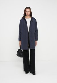 Parka impermeabile blu con cappuccio, dotata di chiusura con zip, vita regolabile con cordoncino e tasche laterali; abbinata a pantaloni neri a gamba larga e una borsa nera.