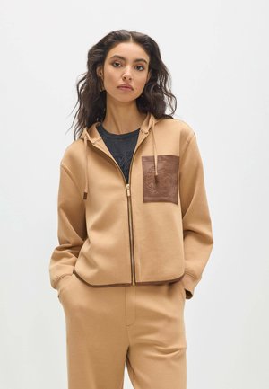 Mujer con cabello oscuro y ondulado, vistiendo una sudadera con capucha beige con cremallera, bolsillo marrón en el pecho y pantalones beige a juego, de pie frente a un fondo blanco liso.