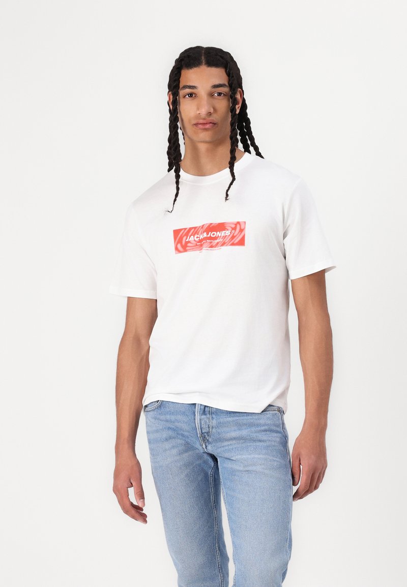 Hvid bomulds t-shirt med et rødt grafisk logo placeret centralt på brystet. Båret med lyseblå denimjeans, der har en afslappet pasform.