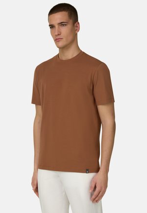 Jeune homme portant un t-shirt à manches courtes marron uni et un pantalon blanc, debout devant un fond clair uni, légèrement tourné vers la gauche.