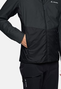 Veste noire à fermeture éclair avec une finition mate contrastante, dotée de poches latérales zippées et de détails de logo. Portée sur un pantalon noir.