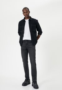 Giubbotto di jeans nero con tasche frontali sopra una t-shirt bianca, abbinato a jeans scuri e scarpe da ginnastica nere con un design testurizzato.