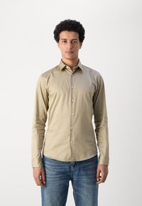 Világos bézs gombos ing hosszú ujjakkal, hegyes gallérral és slim fit szabással. Finom fényű és fehér gombokkal rendelkezik. Kék farmernadrággal viselve.