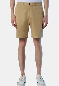 Man in beige knielange shorts en witte sneakers, staand tegen een effen lichte achtergrond.
