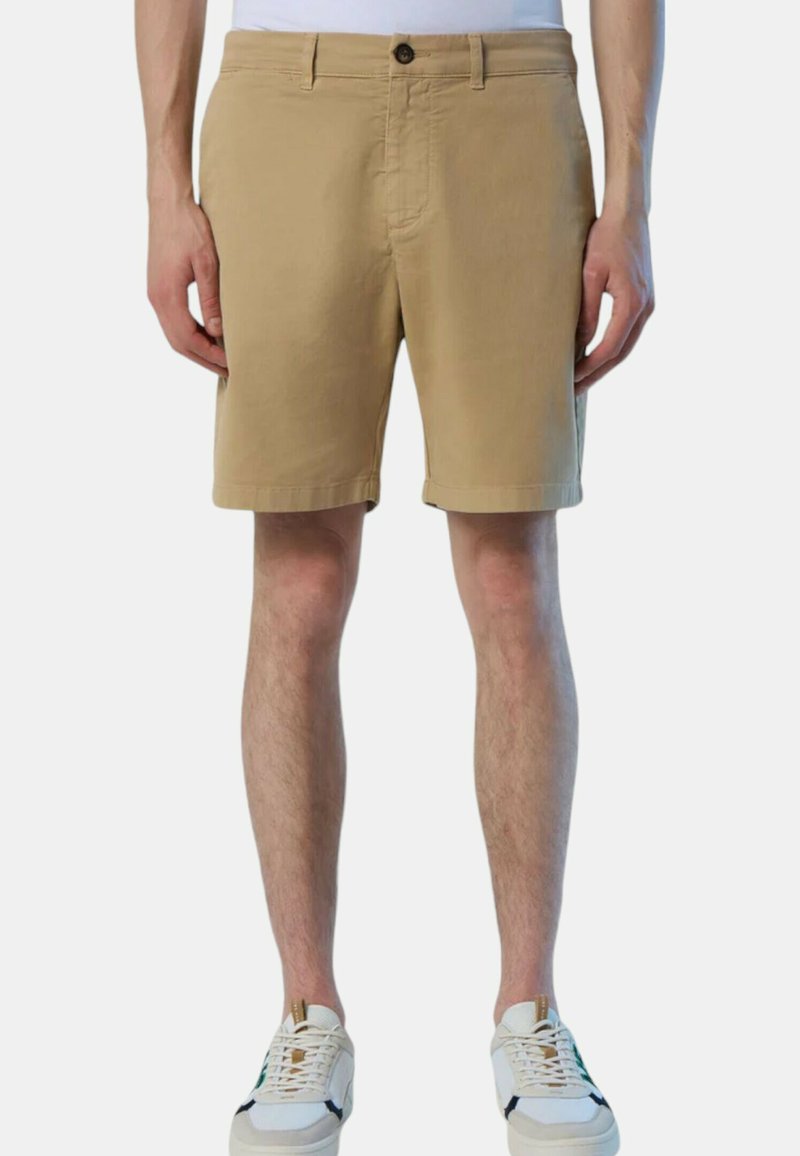 Man in beige knielange shorts en witte sneakers, staand tegen een effen lichte achtergrond.