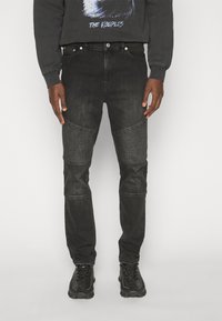 Black denim jeans with textured knee patches, vertical ribbed detailing, and a slim fit design. Paired with black footwear and a dark sweatshirt.