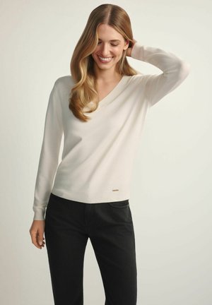 Uśmiechnięta kobieta o długich blond włosach, ubrana w kremowy sweter z dekoltem w serek i czarne spodnie, stojąca na jasnym tle.