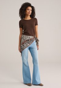 Bruin kortermig t-shirt gecombineerd met lichtblauwe uitlopende jeans. Een patroonrijke bruine en crèmekleurige wikkel is in de taille gestrikt, met ingewikkelde ontwerpen.