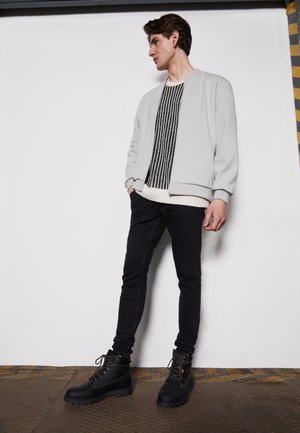 Chaqueta bomber gris sobre un suéter de rayas en blanco y negro, combinada con jeans negros y botas con cordones negras. Texturas suaves, diseño ajustado.
