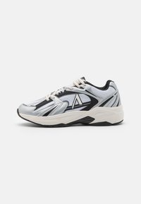 Scarpa sportiva argento e nera con sovrapposizione in rete, caratterizzata da una suola spessa, un logo prominente e lacci a contrasto per supporto e stabilità.