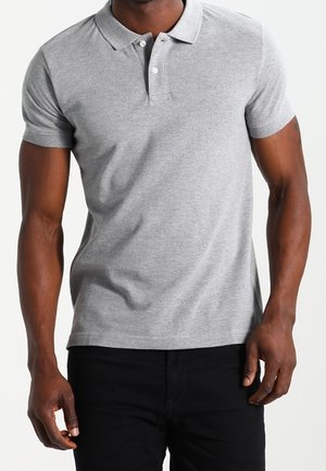 Homme portant un polo gris clair à manches courtes avec une patte de boutonnage à deux boutons et un pantalon noir, debout avec les bras détendus.