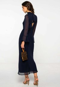 Vestido de manga larga azul marino con mangas transparentes, espalda con corte y silueta ajustada, combinado con un bolso de mano dorado con lentejuelas.