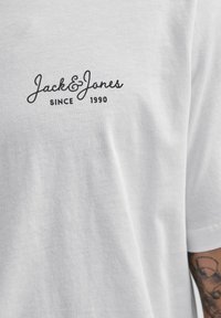 Baltas marškinėliai su juodu tekstu "Jack & Jones Since 1990" ant krūtinės ir daliniu tatuiruoto rankos vaizdu dešinėje pusėje.
