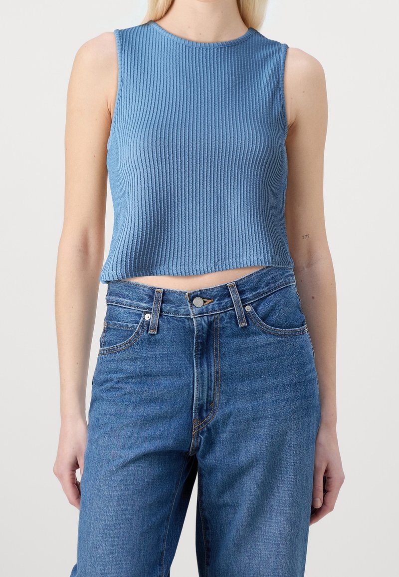 Crop top bleu côtelé sans manches porté avec un jean bleu taille haute sur une personne debout.
