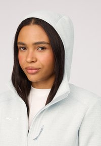 Giacca a vento in fleece azzurro chiaro con zip frontale, texture sottile e dettaglio del logo sul petto. Presenta un cappuccio aderente.