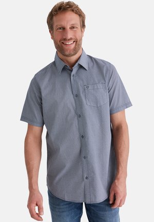 Lächelnder Mann, der ein kurzärmliges, blau gemustertes Hemd mit Knöpfen und Jeans trägt, vor einem schlichten weißen Hintergrund stehend.