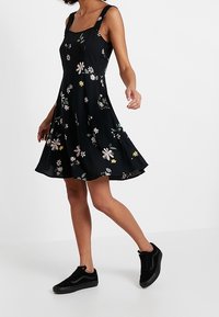 Robe fleurie noire avec un corsage ajusté, une jupe évasée et des bretelles réglables, présentant des motifs floraux multicolores et une texture lisse.