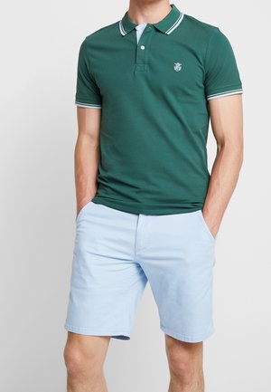 Polo verde con colletto e dettagli delle maniche bianchi, abbinato a pantaloni corti azzurri chiari. Tessuto liscio, design aderente, con un piccolo logo sul petto.