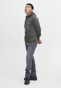 Oliven grøn hoodie med logo og grafik, parret med grå jeans og sorte snørebåndsstøvler. Enkel design, afslappet pasform og blød tekstur.