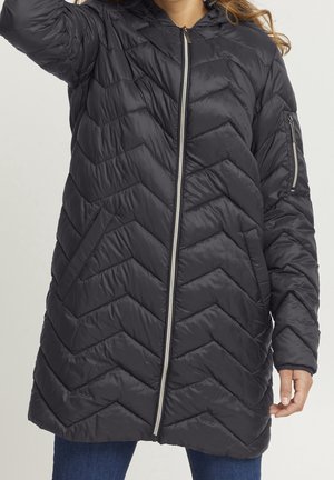 Kobieta ubrana w długą czarną pikowaną kurtkę z przednim zamkiem błyskawicznym, kieszenią na rękawie zapinaną na zamek i częściowo widocznymi rękami.