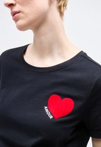 Άτομο που φοράει μαύρο πουκάμισο με ένα κόκκινο καρδιά και τη λέξη «AMOUR» τυπωμένη στο στήθος, απέναντι από φόντο απαλού φωτός.