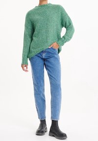 Persona con un suéter de punto verde, jeans azules y botas negras de tacón grueso, de pie con una mano en el bolsillo contra un fondo blanco.