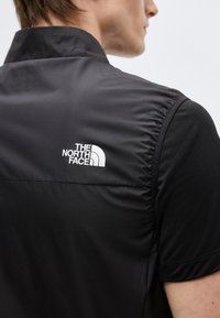 Primo piano di una persona che indossa un gilet nero con il logo The North Face sulla parte superiore della schiena e una maglietta nera a maniche corte sotto.