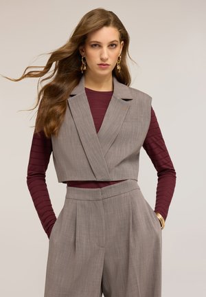 Donna con capelli lunghi e ondulati indossa un blazer grigio senza maniche corto sopra una maglia a maniche lunghe color bordeaux e pantaloni grigi a vita alta abbinati, con le mani in tasca.