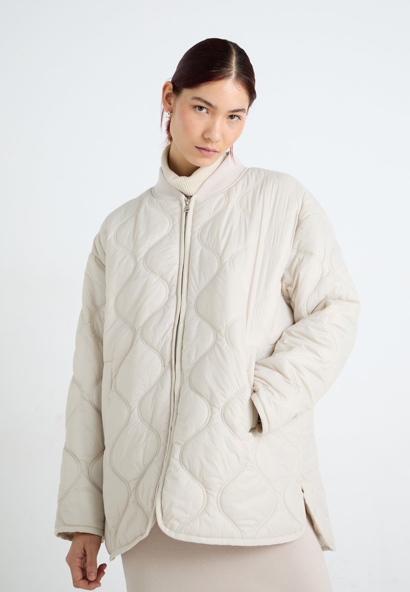 Quiltet, lys beige jakke med høy krage, frontglidelås og sidesplitter. Har et bølgete teksturmønster på stoffet. Mykt, polstret materiale.