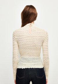 Langærmet top i creme med teksturerede striber og en nøglehul-detail i ryggen. Blødt stof med en tætsiddende silhuet og bølget kant.