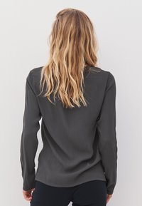 Woman with long wavy blonde hair wearing a dark gray long-sleeve top and black pants, facing a plain white wall.