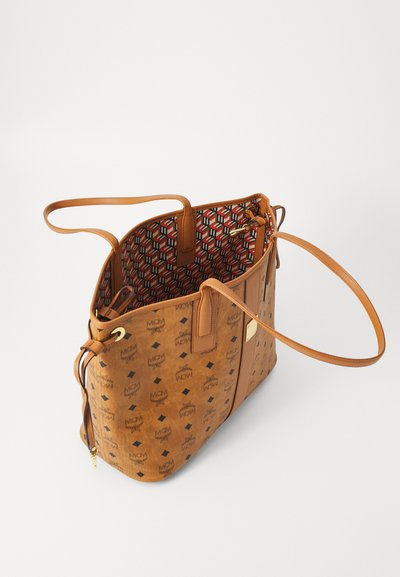 Brown leather tote bag with MCM logo pattern, long handles, and geometric red and black inner lining, open at the top.