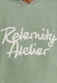 Lichtgroene gebreide trui met een V-hals ontwerp, voorzien van witte tekst die "Referenity Atelier" leest.