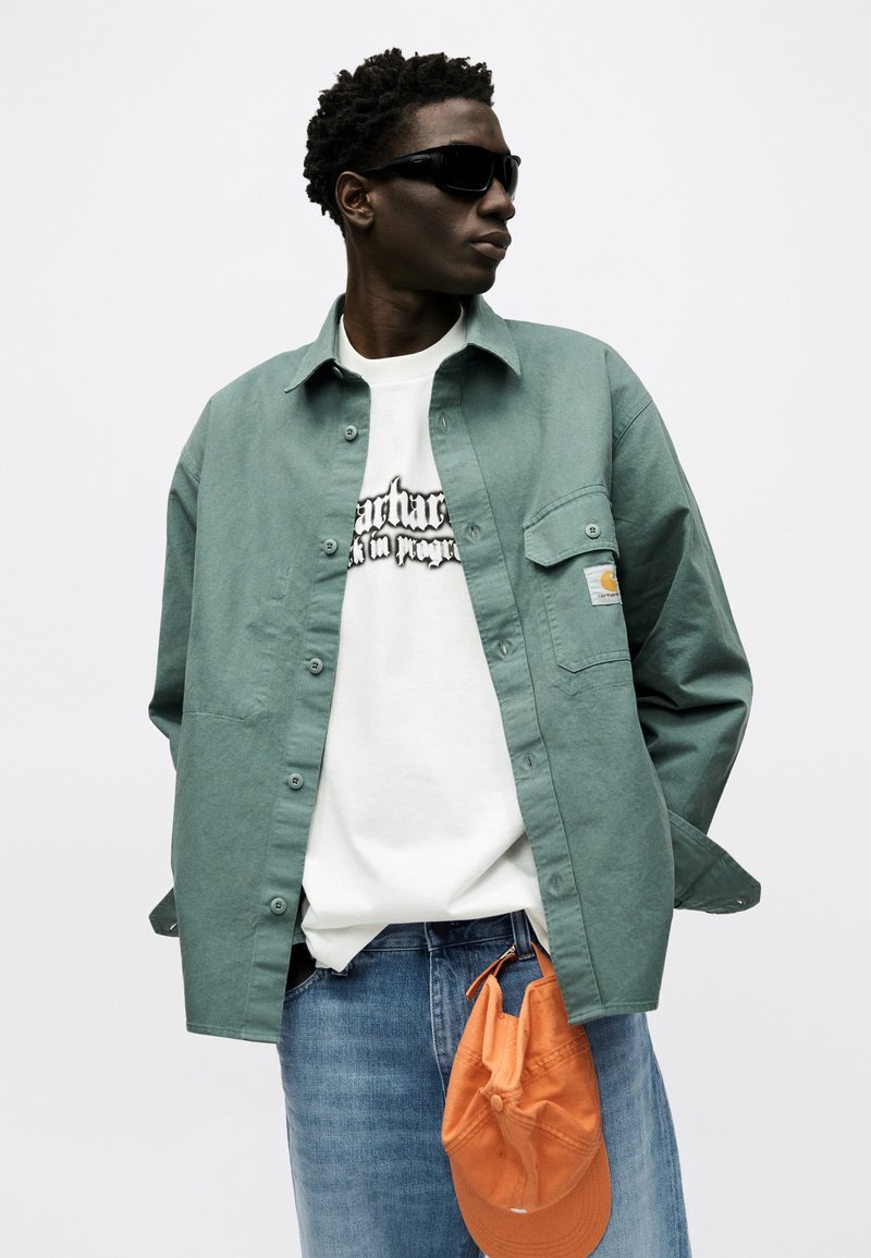Young man wearing black sunglasses, green overshirt, white graphic tee, blue jeans, holding an orange cap against a white background.